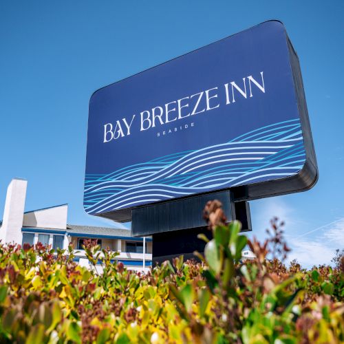 A large blue sign reads &ldquo;Bay Breeze Inn&rdquo; with wavy white lines beneath; some shrubs are in the foreground and a building is in the background.