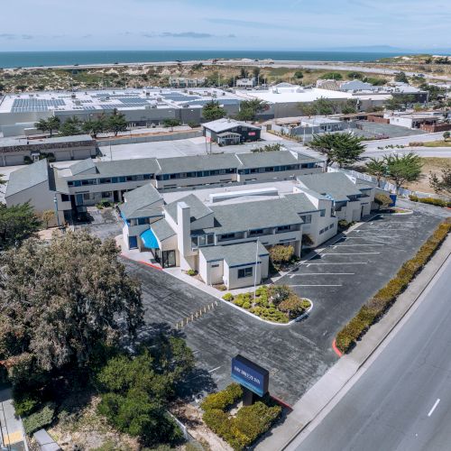 Aerial view of a modern commercial complex beside a busy road, with parking lots, low-rise buildings, blue accents, and surrounding desert landscape.