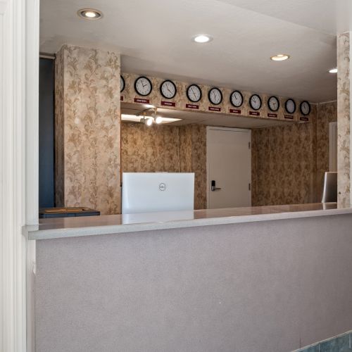 A reception or front desk area with a long counter, beige walls, a row of clocks on the back wall, and a doorway behind the counter.