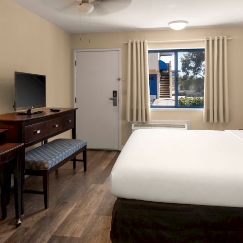 A clean hotel room with a big bed, desk, TV, and window view, in neutral tones with a red accent wall, ready for guests.