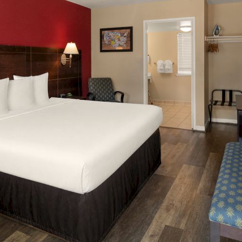 A hotel room with a large bed, red accent wall, two nightstands, a desk, chair, bench, and open closet in the back.