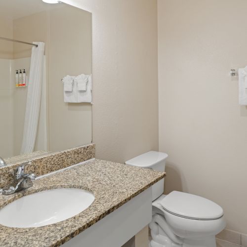 A clean bathroom with a granite countertop sink, mirror, toilet, and a towel rack with folded towels on the wall.
