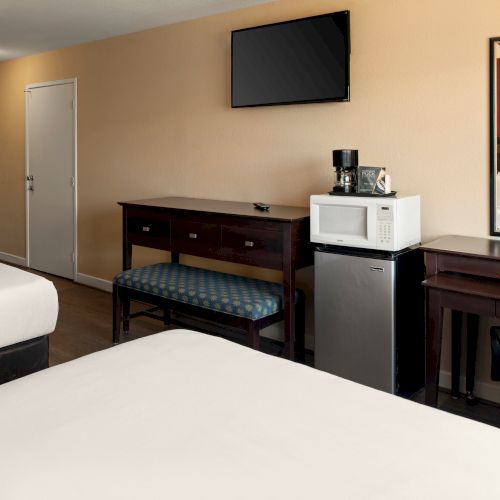 A hotel room with two beds, a wall-mounted TV, a desk and chair, a mini-fridge with a microwave, and a dresser; coffee setup near a mirror.