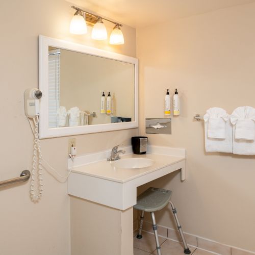 A small beige bathroom with a sink, wall mirror, hairdryer, wall-mounted shelves, two folded towels, a stool, and a window beside the sink.