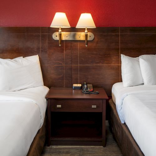 Two neatly made beds with white linens, a shared nightstand, wall lamps, and a red-accented headboard wall.