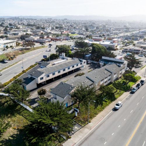 An aerial view of a suburban area with streets, buildings, and parked cars, surrounded by trees and greenery under a mostly clear sky.