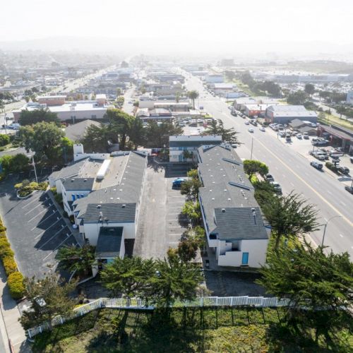 Aerial view showing a row of buildings along a major street with light traffic and greenery, in an urban area under a clear sky.