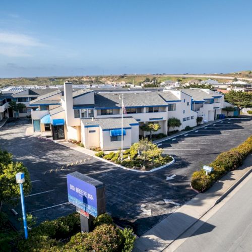 An aerial view of a building complex with a large parking lot, surrounded by greenery and roads, under a clear blue sky.