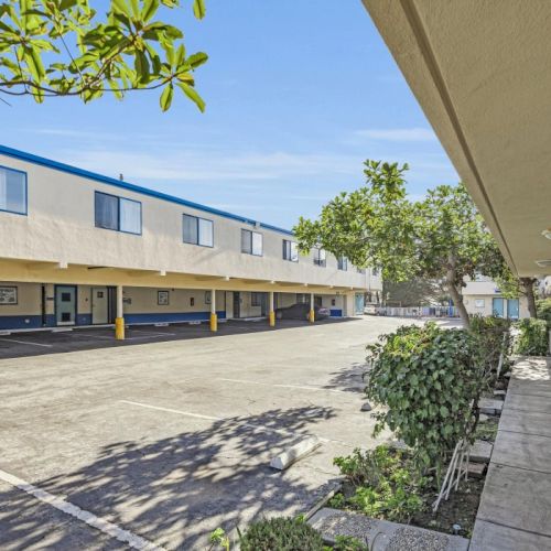 A motel with parked cars, two-story building, blue doors, and outdoor passageways surrounded by trees and shrubs.