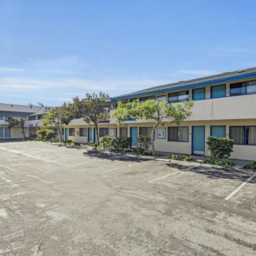 The image shows a two-story motel with a parking lot, lined with small trees and shrubs, under a clear blue sky.