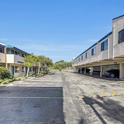 A paved area between two sets of multi-story buildings with carports, plants, and parked vehicles under a clear blue sky.