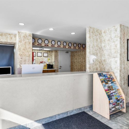 The image shows a hotel reception with a counter, brochure stand, a table with chairs, and decorated walls.