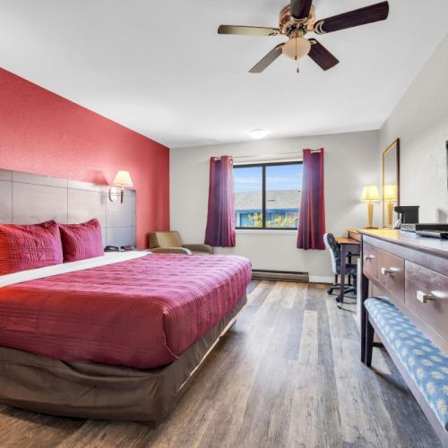 A hotel room with a large bed, TV on a dresser, wall-mounted lamps, a window view, ceiling fan, and wood flooring.