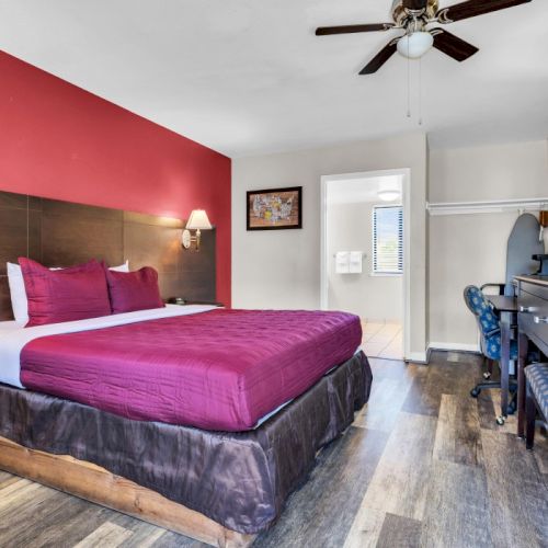 The image shows a hotel room with a red-accented wall, bed, lamps, TV, and a desk with a chair. There's also a bathroom entrance visible.