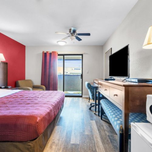 A hotel room featuring a double bed, flat-screen TV, desk, chair, microwave, and lamp, with a sliding door to a balcony.