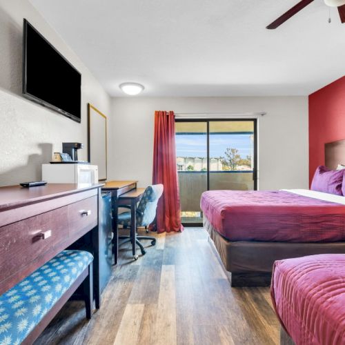 A hotel room with two beds, red accents, a TV, desk, and window with curtains, leading to a balcony with a view outside.