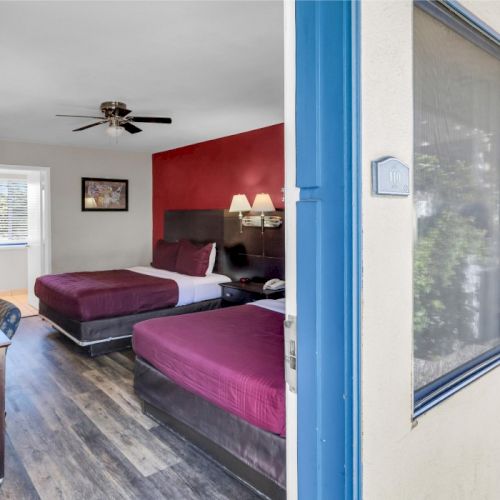 The image shows a hotel room with two beds, red walls, a desk, and a window, viewed from an open door outside.