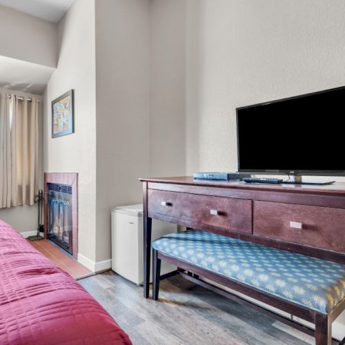 The image shows a hotel room with a bed, dresser, TV, mini-fridge, a bench, fireplace, and windows with curtains.