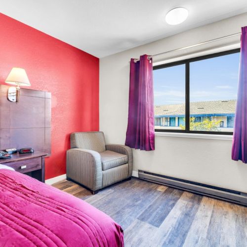 A cozy room with a red accent wall, bed, armchair, lamp, window with purple curtains, and wooden floor, creating a warm atmosphere.