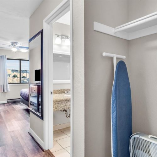 A hallway leads to a room with a bed, TV, desk, and chair. An ironing board and a fan are in a closet area.