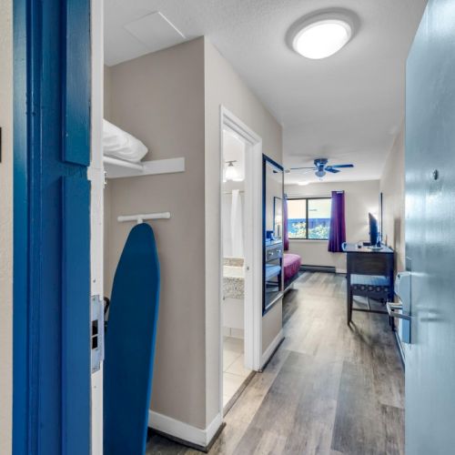 An open hotel room door reveals a room with an ironing board, a bed, and a view of the outside through a window.