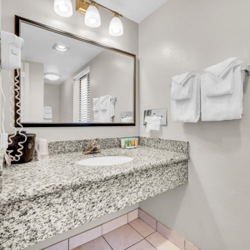 The image shows a modern bathroom with a granite countertop, sink, mirror, light fixtures, hairdryer, and neatly folded towels on the wall.
