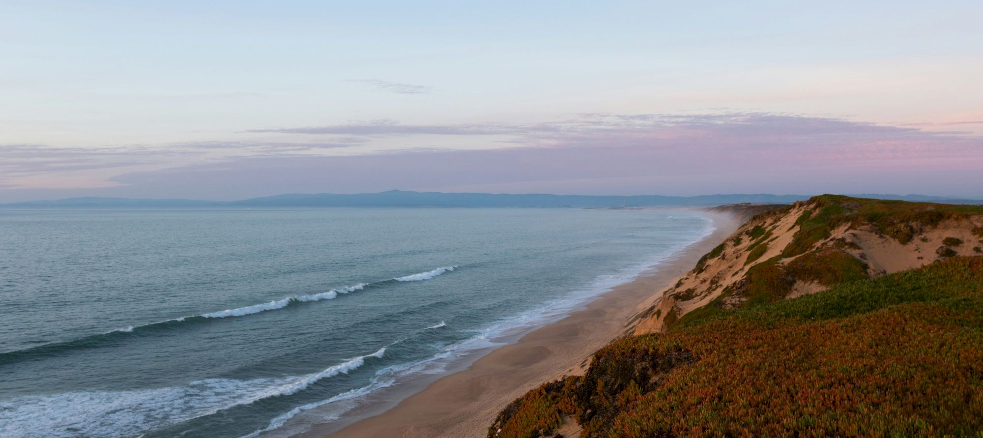 A scenic coastal landscape with waves gently lapping the shore and lush vegetation covering the sand dunes under a serene sky.